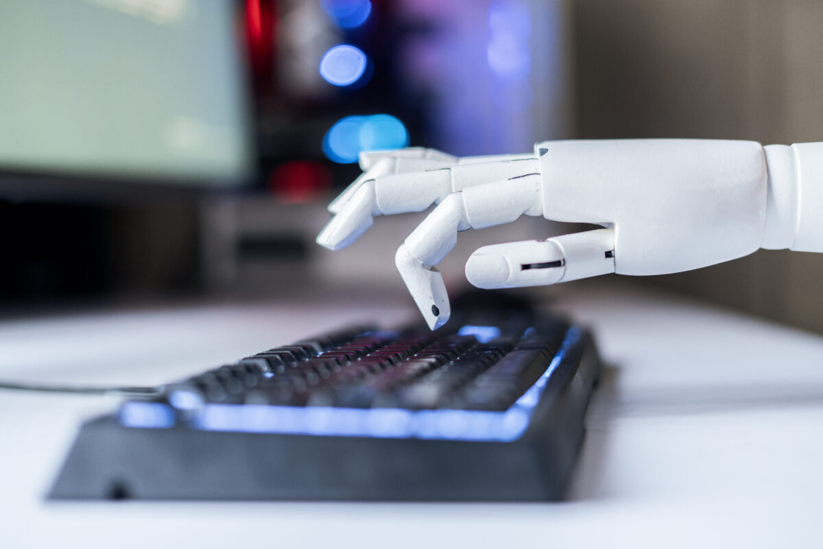 Close-up of a white robotic hand reaching to type on a backlit computer keyboard with a blurred monitor and colorful lights in the background