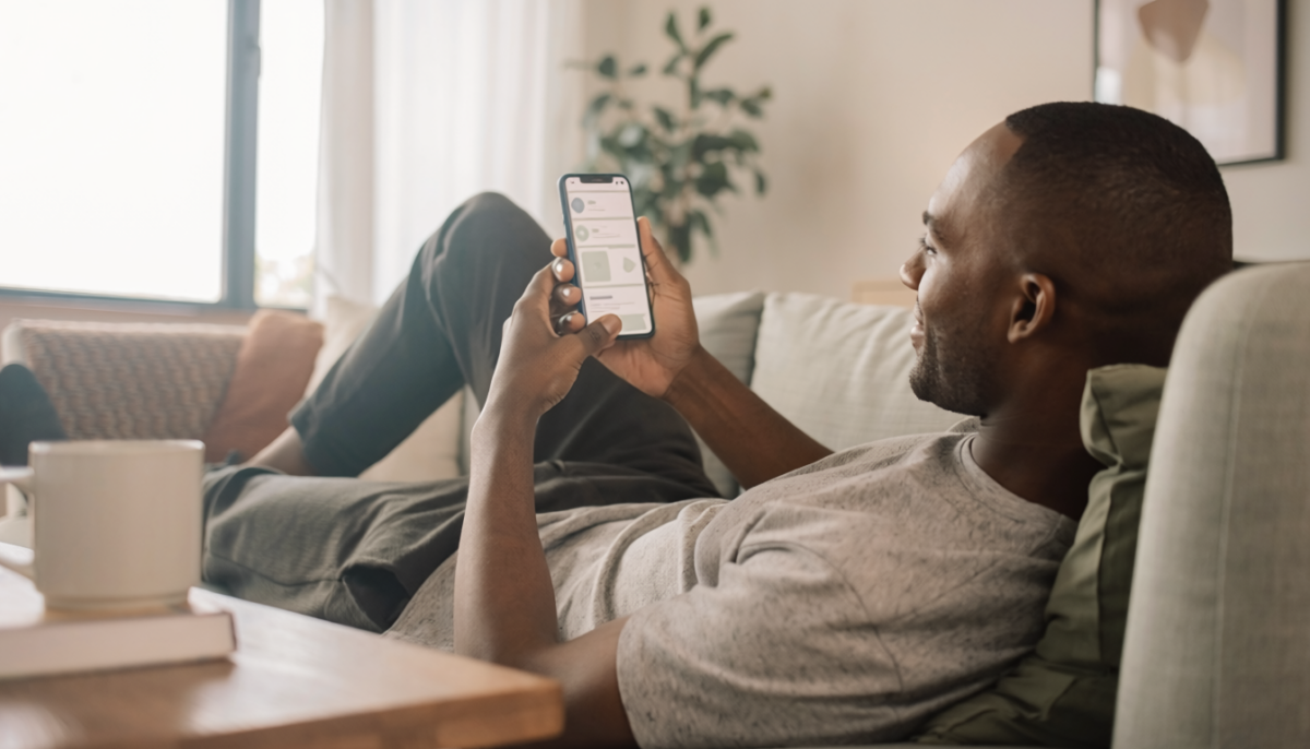 Man relaxing on a couch at home while using a smartphone to browse a new mobile app, with a coffee mug on a nearby table and soft natural light coming through the window.